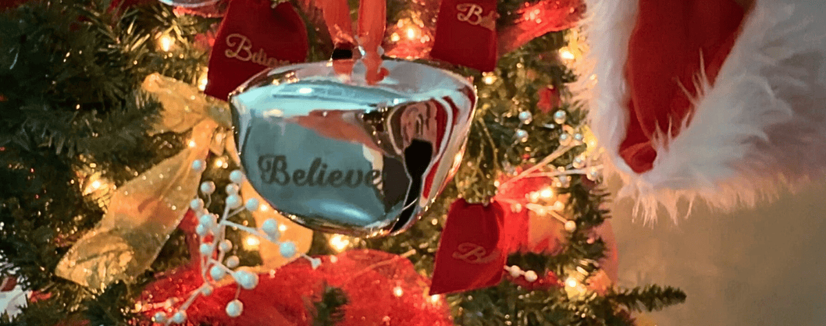A silver bell with the word "believe" on it hands in front of a Christmas tree.