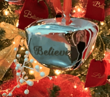 A silver bell with the word "believe" on it hands in front of a Christmas tree.