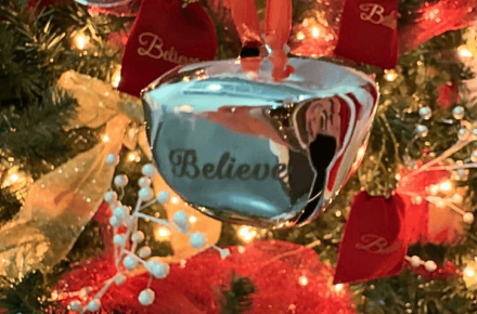 A silver bell with the word "believe" on it hands in front of a Christmas tree.